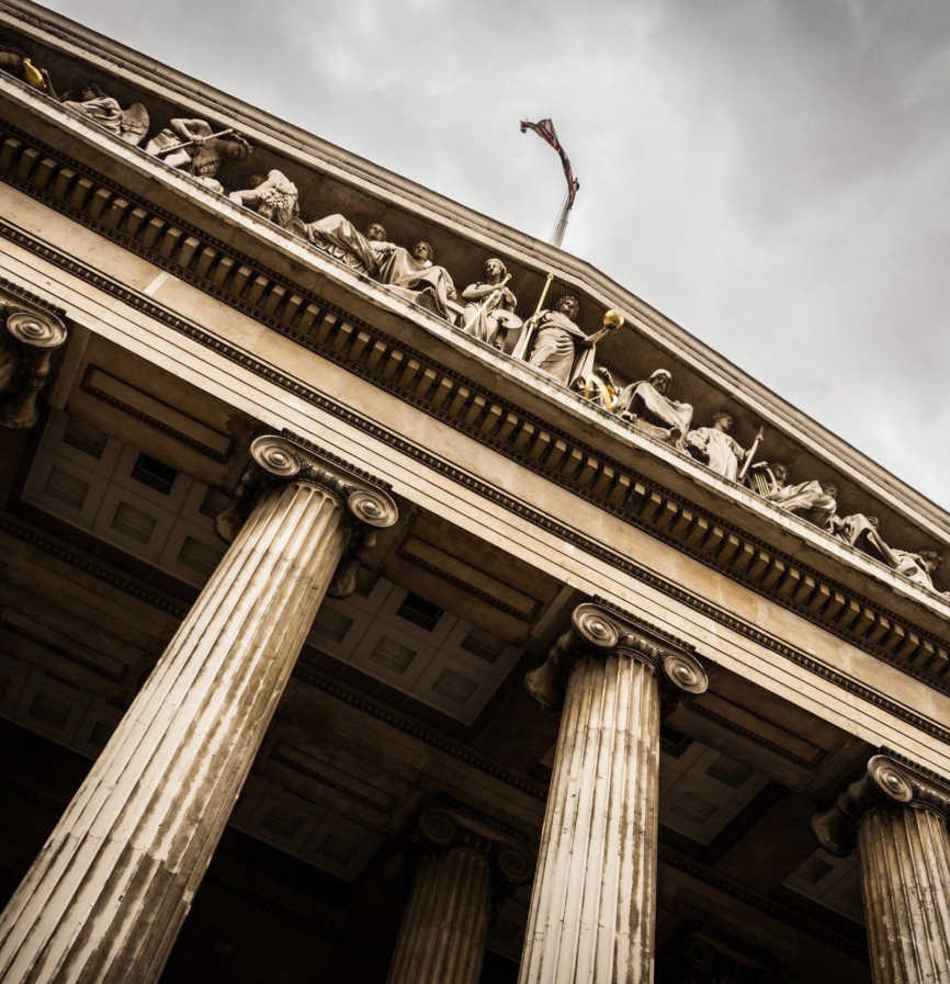 Classical building facade with tall columns, sculptural frieze, and flag against cloudy sky